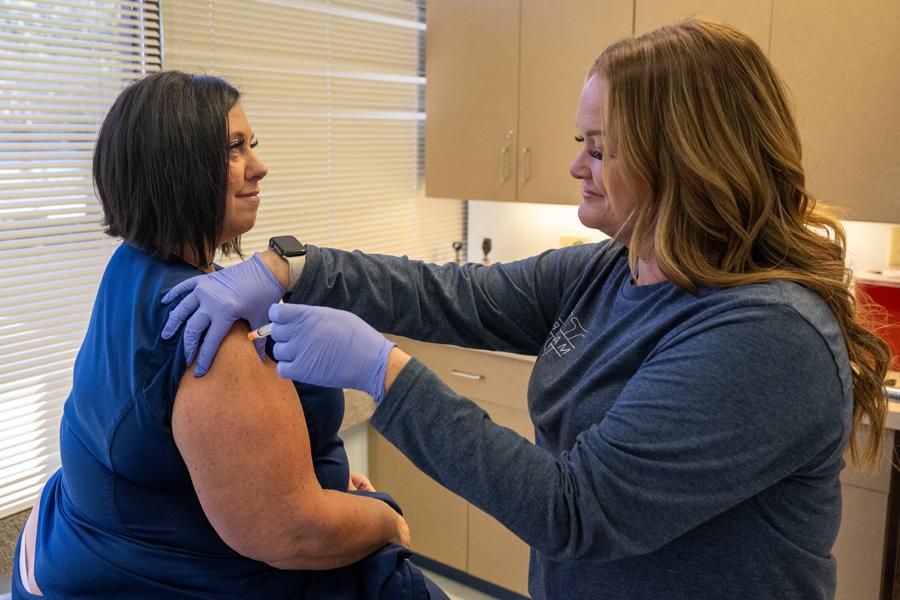 A smiling woman with dark blonde hair wearing a gray sweater and blue lab gloves administers an injection to a woman with short, dark brown hair wearing a blue shirt with a sleeve rolled up. They are standing in an exam room in front of a window with closed blinds. White cabinets are visible behind them.