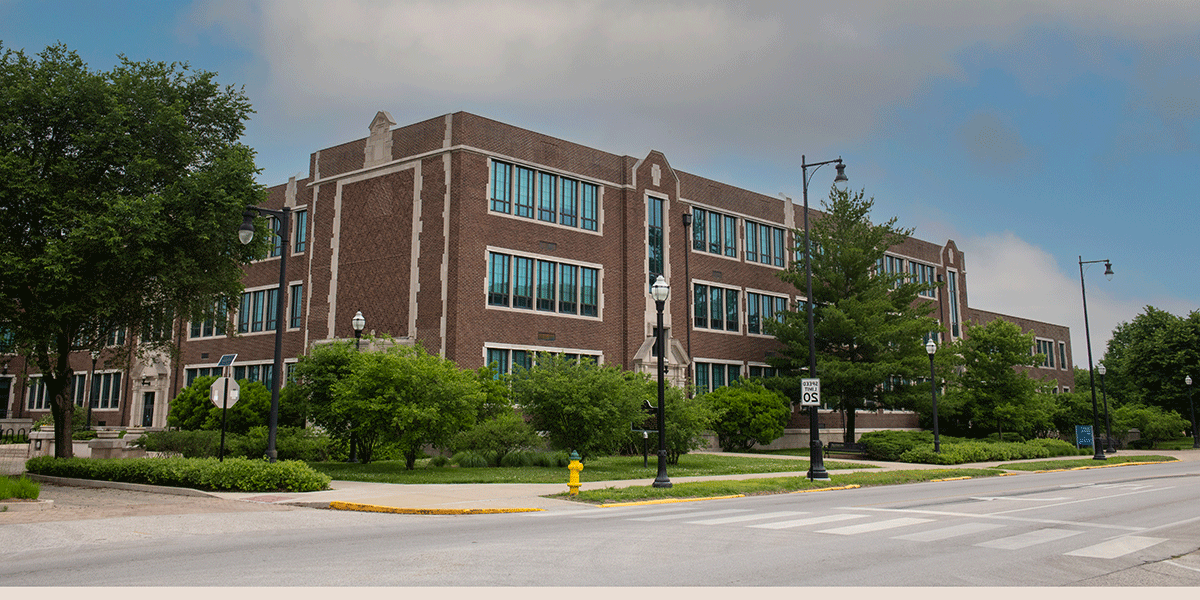 A red brick building with white trim, as viewed from the street. A lamppost, trees and greenery, and a speed limit sign showing 20 mph are all visible in front of the building. Blue sky and greyish-white clouds are visible above.