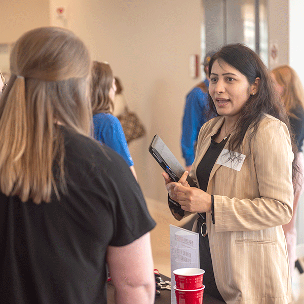 A woman talking to another woman. They are standing at a table and seem to be talking to one another. 