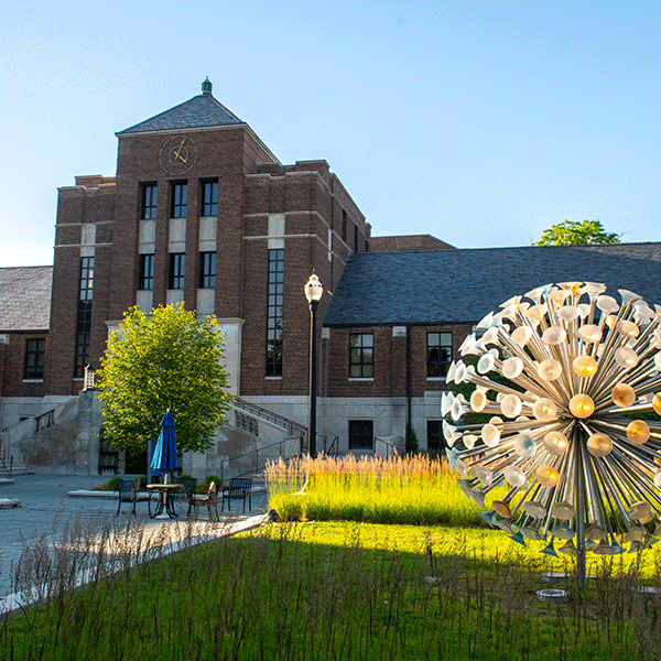 A large brick building with a large clock with a large, circular art installation in front with green grass.     