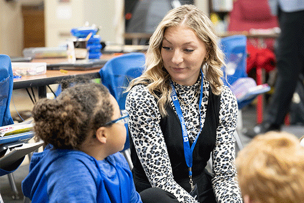 A white female student with shoulder-length curly blond hair sits in a classroom. She wears a cheetah-print, black-and-white, long-sleeved shirt, a black vest, and a blue lanyard. On her right is a young Black female child with brown hair pulled back into a bun. She wears a blue hoodie. Another child’s blond hair is visible to her left, and tables and blue chairs are visible in the background. 