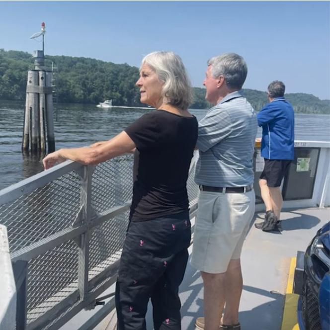 The image shows a ferry deck with two individuals: a woman in a black shirt and pants and a man in a light blue shirt and white shorts, both leaning on the metal railing. The background features a navigational buoy and green trees along the water's edge, with a sunny atmosphere and clear visibility.