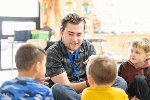 A white, male Indiana State student-teacher, with brown hair and wearing a gray camo t-shirt and blue lanyard, sits and talks with two young boys in a classroom. He is smiling.