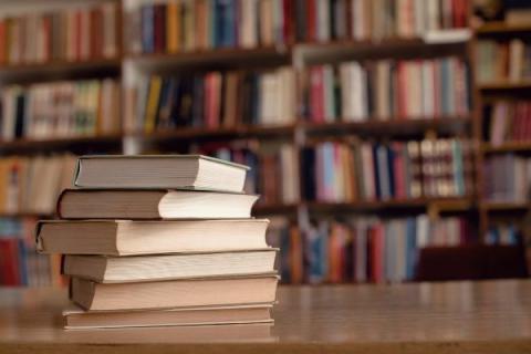 A stack of six books sits on a table in the library. The spines are facing away and cannot be read. In the background, shelves of colorful books are visible.