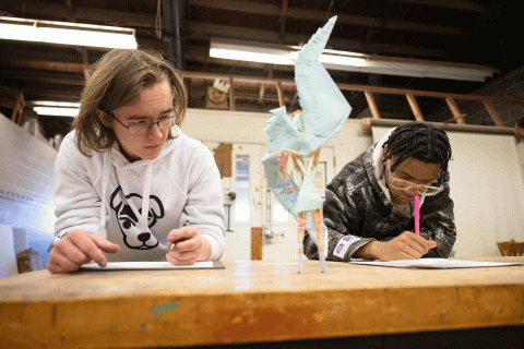 Two students seated in a workshop observe a sculpture that is on a table while writing their observations on paper.