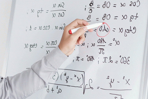 A close-up picture of a whiteboard with multiple equations written on the board in black lettering. A white individual’s hand is holding a red dry erase marker to the whiteboard, circling an equation. The person is wearing a long-sleeved white dress shirt.   