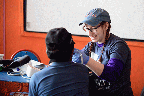 A white female student with a denim hat and glasses wearing blue surgical gloves holds an instrument towards a young patient with his back turned to the camera in a rural South American clinic with orange walls and a whiteboard in the background. 