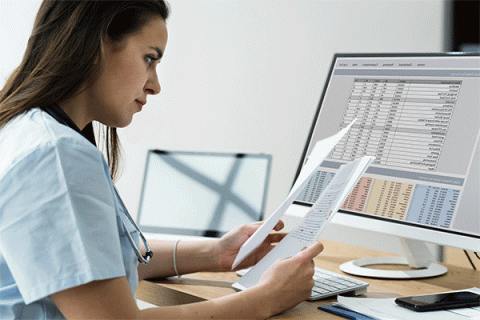 Female wearing scrubs and seated at a desk reviewing papers and a spreadsheet on a desktop in the background. 