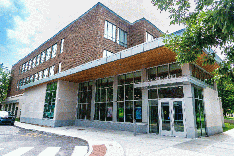 Exterior view of Dreiser Hall, a modern brick-and-glass building with large windows and a wooden overhang at the entrance. The entrance features double glass doors with the name 'Dreiser Hall' above them. There are trees and a car visible on the side, with a bright sky in the background.