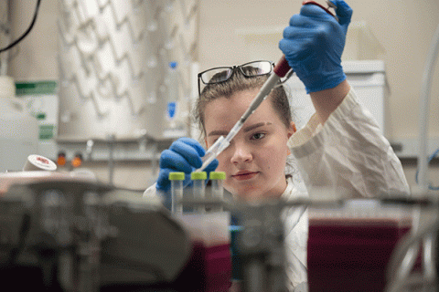 A female student with dark blonde hair, wearing a white lab coat, and with brown glasses pushed up onto her head, faces the camera as she uses a pipette in a lab setting. Various beakers and pieces of equipment are blurred but visible on a shelf in the foreground. 