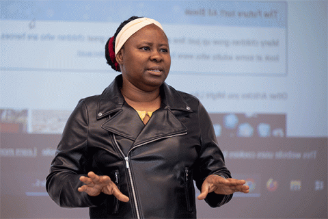 Black female professor wearing a black leather jacket and a white headscarf stands in front of an illuminated screen displaying a web page as she speaks to a class.