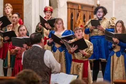 A group of singers in medieval-style costumes performs together. They hold music sheets and display various expressions of concentration and joy. The background features ornate wooden architecture, enhancing the festive atmosphere.